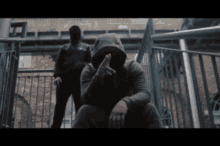 a man in a hooded sweatshirt is sitting on a railing with another man standing behind him