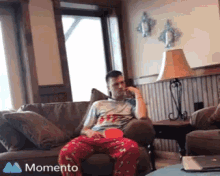 a man in pajamas is sitting on a couch with a momento icon in the corner