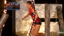 a woman in a red tank top is jumping over a barrier with a scoreboard that says titanes 6 5 heroes