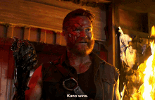 a man with blood on his face is standing in front of a fire and says kano wins