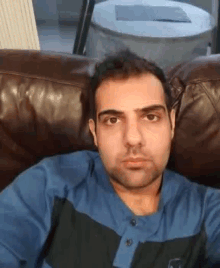a man in a blue shirt is sitting on a brown leather couch .