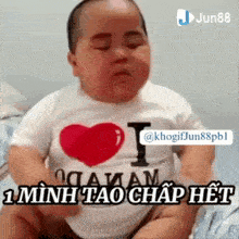 a baby is sitting on a bed with his eyes closed and wearing a t-shirt with a heart on it .