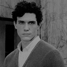 a black and white photo of a man with curly hair wearing a sweater and a striped shirt .