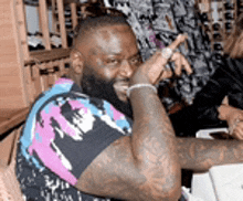 a man with a beard and tattoos is sitting at a table with his hand on his face .