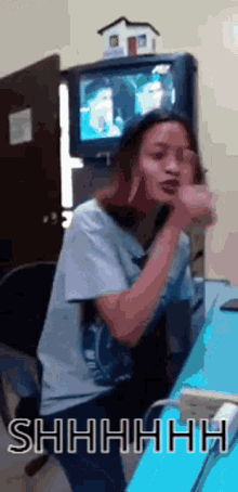 a woman is sitting at a desk in front of a television and making a shhh gesture
