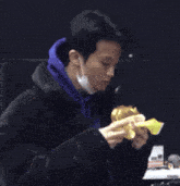a man wearing a blue hoodie and a black jacket is eating a sandwich