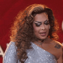 a drag queen with a tattoo on her arm is wearing a silver dress and making a funny face .