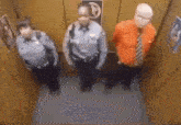 three police officers are standing in an elevator with a man in an orange shirt .