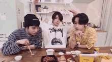 three young men are sitting at a table eating food . one of them is wearing a stussy shirt .