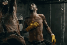 a shirtless man wearing yellow rubber gloves is standing in front of a large piece of meat