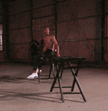 a shirtless man sits on a folding chair in a warehouse