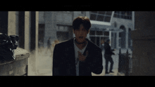 a man in a suit and choker stands in a dark alleyway with a trash can that says ' trash ' on it