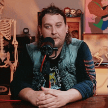 a man in a denim vest is sitting in front of a microphone with a skeleton in the background