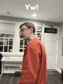 a man in an orange sweater is standing in a living room with a keyboard in the background and the words yw written on the bottom right
