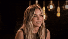 a woman with long blonde hair is smiling in a dark room with light bulbs hanging from the ceiling .