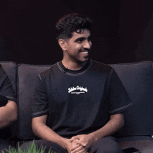 a man sitting on a couch wearing a black t-shirt that says ' hidden original ' on it