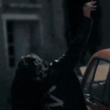 a woman in a black jacket is standing next to a red car with her arm in the air .