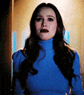 a woman wearing a blue turtleneck sweater and red lipstick is standing in a dark room .