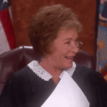 a judge with glasses and a lace collar smiles while holding a piece of paper