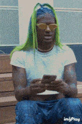 a man with green hair and yellow sunglasses is sitting on a bench looking at his phone