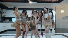 a group of women dancing in a kitchen with one wearing a plaid skirt