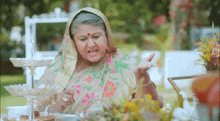 an elderly woman in a sari is sitting at a table with flowers and drinking tea .