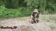 a man wearing a hat is kneeling on a rocky river bank .