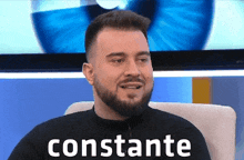 a man with a beard is sitting in front of a television and the word constante is on the screen