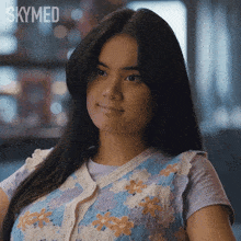 a woman is wearing a blue and white sweater vest with flowers on it and a skymed logo behind her