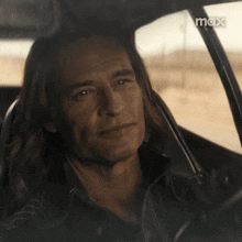 a man with long hair is smiling in a car with the word max on the bottom right