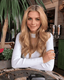 a woman with long blonde hair is sitting at a table with her arms crossed