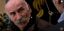 an older man with a mustache is talking to another man in front of bananas