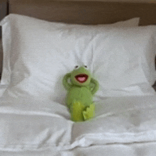a stuffed kermit the frog is sitting on a bed with white sheets .