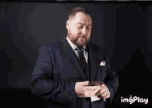 a man in a suit and tie is holding a cell phone in front of a black background that says imgplay on it