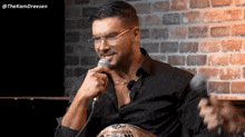 a man wearing glasses and a black shirt is holding a microphone in front of a brick wall ..