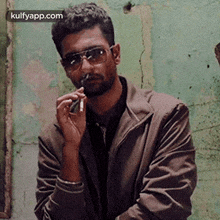 a man wearing sunglasses and a jacket is smoking a cigarette in front of a green wall .