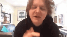 a man in a black hoodie is sitting in a living room with his eyes closed and his hands on his chest .