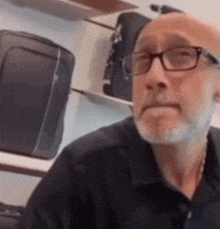 a bald man with glasses and a beard is sitting in front of a shelf of luggage .