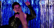 a man in a leather jacket is singing into a microphone while dancing in front of a blue curtain .