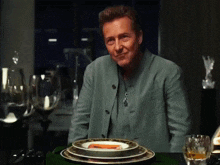 a man in a green jacket sits at a table with plates and wine glasses