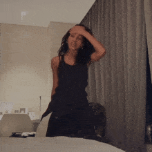 a woman in a black tank top holds her hand to her forehead in a room
