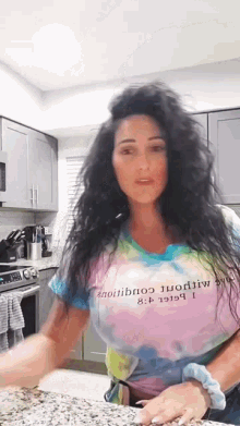 a woman with long hair is standing in a kitchen wearing a tie dye shirt .