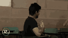 a man in a black shirt is sitting at a desk in a classroom and making a funny face