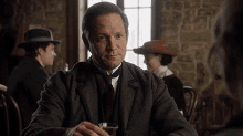 a man in a suit and tie is sitting at a table with a glass of whiskey in his hand .