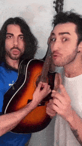 a man in a blue puma shirt holds a guitar next to another man in a white shirt