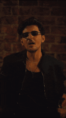 a man wearing sunglasses and a black jacket is standing in front of a brick wall in a dark room