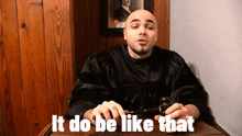 a bald man sitting at a table with the words " it do be like that " written on the bottom