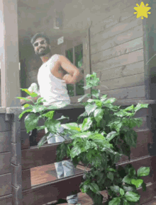a man in a white tank top stands on a porch next to a plant