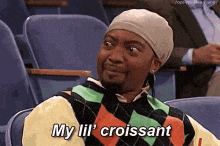 a man wearing a hat and sweater is sitting in a chair and saying my lil croissant .