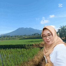 a woman wearing glasses and a hijab stands in front of a lush green field
